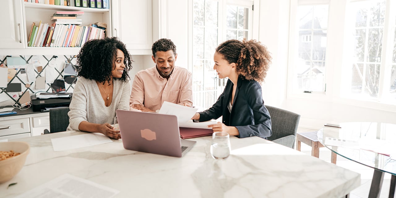 Photo of three adults sitting at a kitchen table reviewing papers and looking at an open laptop.